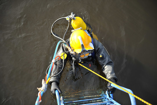 An industrial diver with scuba diving and in a protective suit climbs out of the water along the stairs.