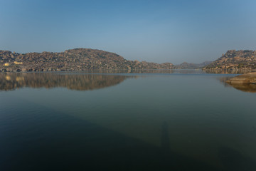 Fototapeta premium Lake in Hampi. Reflections in the water of the cave city and rocks in the early morning in the Indian state of Karnataka.A hazy haze can be seen on the horizon.