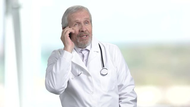 Elderly doctor talking on phone. Senior doctor in white coat talking on mobile phone, abstract blurred background.
