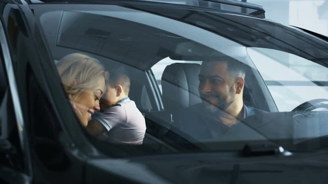 View through front glass of happy kissing parents and chatting with content children sitting in new bought car in showroom. Movement stabilized 4k shot.