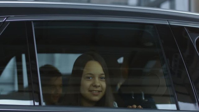 View through glass of charming smiling girl looking at camera sitting with family inside of new car in dealer salon. Movement stabilized 4k shot.