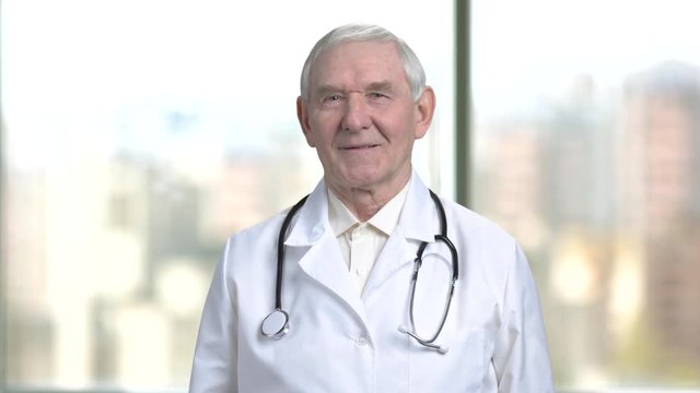 Older medical specialis portrait. Smiling doctor with stethoscope in blurred city view from windows background.