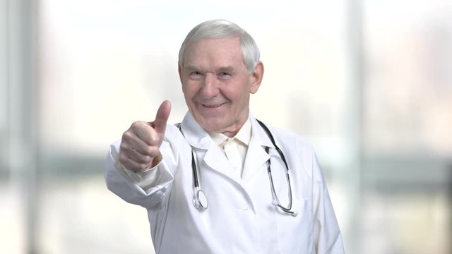 Senior old doctor in white coat and stethoscope shows thumb up. Fist with thumb up turning rotation, blurred windows background.