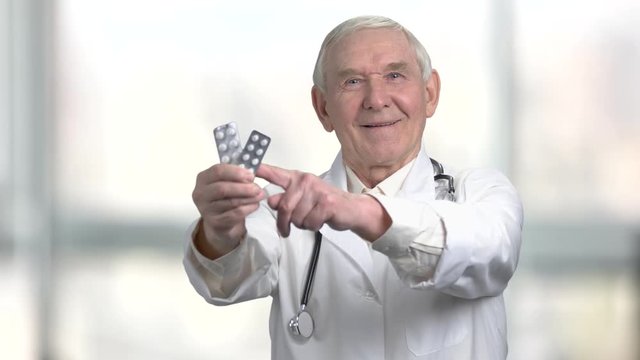Pointing at the pack of medicine pills. Smiling very old doctor recommed pills to you in hospital, bright blurred background.