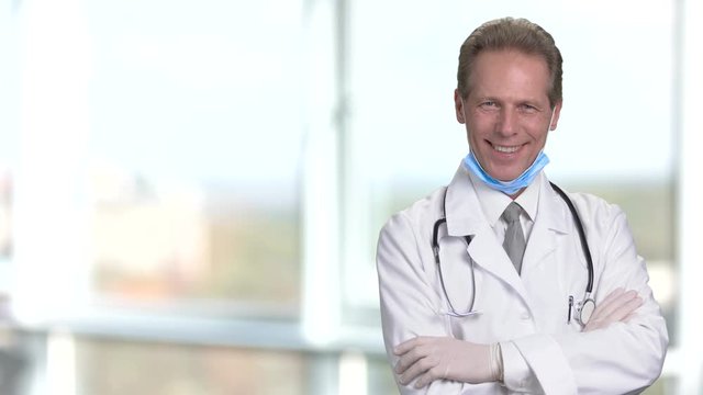 Doctor in white uniform crossing arms. Mature man working as doctor in hospital. Abstract blurred background in bright room.
