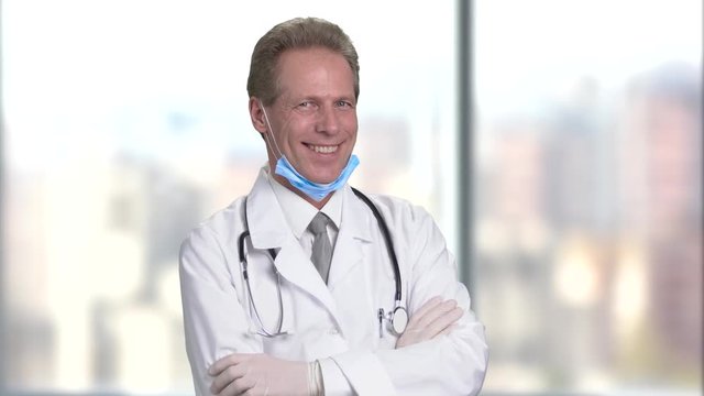 Happy mature middle aged smiling doctor with folded arms, front view. Bright abstract blurred windows background with view on city.