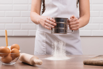 Young woman sifting flour through a sieve on  wooden table. The chef in white apron sifts the flour through a sieve to prepare the dough for pizza on a light background.