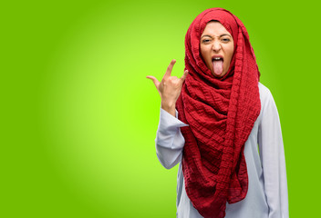 Young arab woman wearing hijab making rock symbol with hands, shouting and celebrating with tongue out