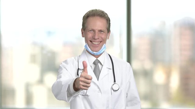 Medical doctor showing thumb up gesture. Smiling man doctor with protective mask under chin showing thumb up, blurred background.
