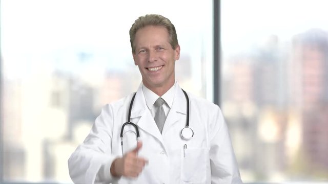 Male doctor putting on stethoscope. Happy doctor cardiologist showing thumb up gesture on blurred background.