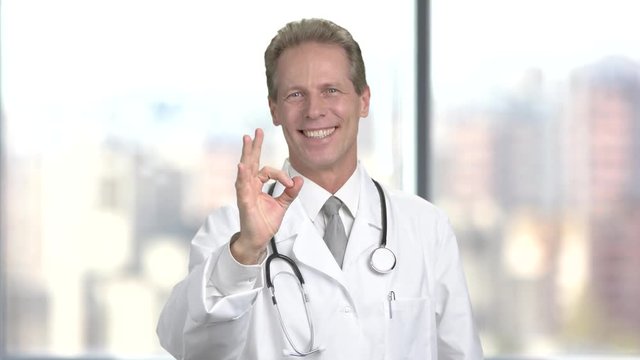 Clinician doctor man showing ok sign. Male smiling physician making sign ok with fingers on blurred background. Success in medicine.