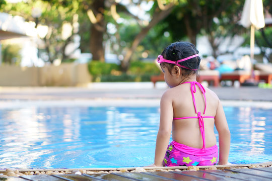 Asian children cute or kid girl backside wear goggles and bikini sit on swimming pool with happy fun in water park for refreshing and relax with exercise on summer holiday travel at hotel with space