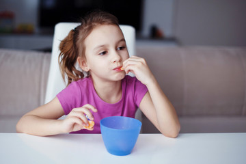 A little girl is sitting at a white table. He drinks from a blue cup and eats cookies.
