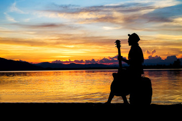 Obraz premium woman traveler have baggage and guitar sitting and looking for view of lake mountain and sunset near the dam on vacation.silhouette concept. 