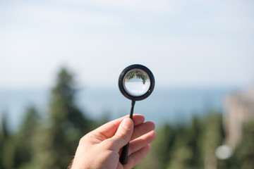 Man hand, magnifying glass in focus, nature, sea, trees on background.
