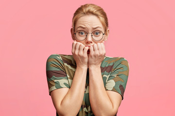 Nervous blonde female student bites finger nails as worries before passing exam, has frustrated look at camera, forgets all necessary information, poses against pink studio background. Excitable woman