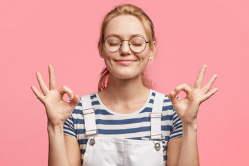 Portrait of satisfied blonde attractive delighted female indicates ok sign, keeps eyes shut, being...