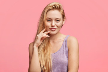 Satisfied mysterious blonde blue eyed young woman has delighted expression, has luxurious light hair, wears casual purple t shirt, thinks about something pleasant, looks directly into camera