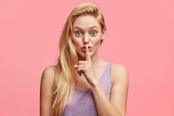 Naklejka premium Beautiful young female blonde young woman keeps fore finger on lips, tells confidential information, demands complete silence, poses against pink studio background. People and secrecy concept