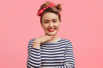 Obraz na płótnie Canvas Candid shot of pleasant looking female with make up, has positive expression, keeps hand under chin, wears striped sweater and stylish headband, poses against pink background. Happiness and people