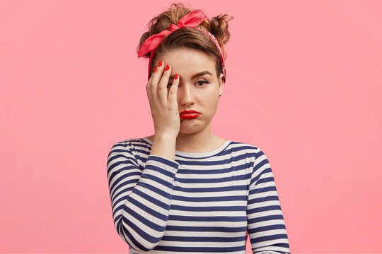 Abused offended discontent beautiful pinup girl wears striped poloneck sweater, red stylish headband, covers face with hand, pouts lips, has retro style in clothing, isolated over pink background