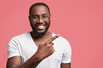 Attractive positive dark skinned man with broad smile, indicates at blank copy space of pink background for your advertising content or promotional text. Happy unshaven male seller of goods.