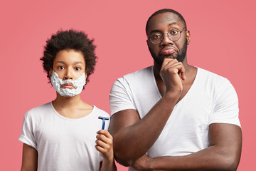 Fatherhood, skin care and hygiene concept. Handsome male teenager learns to shave, has shaving gel on face, holds razor and looks with serious expression, stands near dad who helps and teaches him