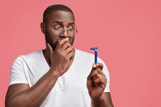 Shocked discontent dark skinned young male looks with unpleasant expression at little razor, can`t imagine how shave thick beard without cream, needs help of barber. African American hairdresser