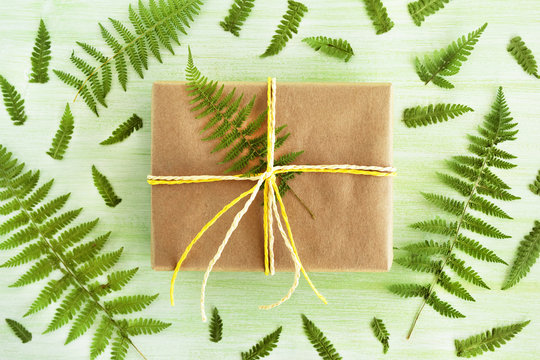 Gift box wrapped of craft paper and white and yellow ribbon on a green wooden background decorated of fern leaves, top view.