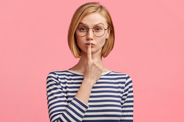 Lovely serious young female model gestures in studio, wears round glasses and striped sailor sweater, demonstrates hush sign, asks to be quiet and not tell secret, poses against pink background