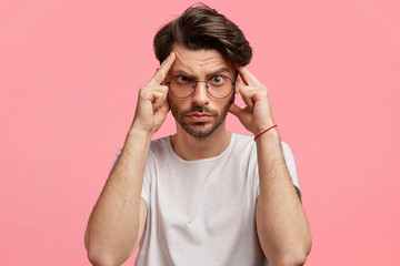 Serious handsome guy tries to concentrate in noisy atmosphere, keeps forefingers on temples, looks with frustrated, tense look, feels tired after hard day, poses against pink studio background