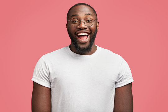 Waist up portrait of glad dark skinned handsome male model with happy expression, wears round spectacles, being in good mood as recieve bonus for diligent work, isolated over pink background