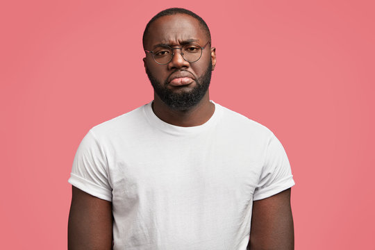 Horizontal studio shot of dissatisfied African American male has gloomy sullen expression, feels tired after hard work at office, being discontent with something, isolated over pink background