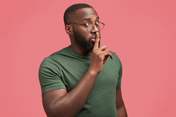 Secret man with dark skin, makes silence gesture, says: Hush, stop talking, looks seriously at camera, wears round spectacles and dark green t shirt isolated over pink background. African guy gestures - obrazy, fototapety, plakaty