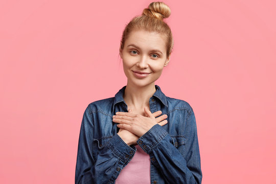 Photo of friendly looking beautiful female being content and touched by something, expresses thankfulness, looks directly at camera with delighted expression, wears fashionable denim jacket.