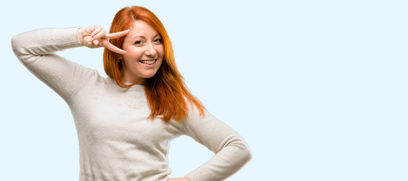 Beautiful young redhead woman looking at camera through fingers isolated over blue background