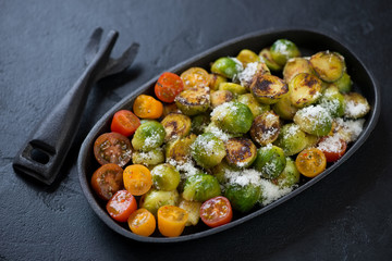Cast-iron pan with roasted brussels sprouts, cherry tomatoes and parmesan over black stone background, studio shot