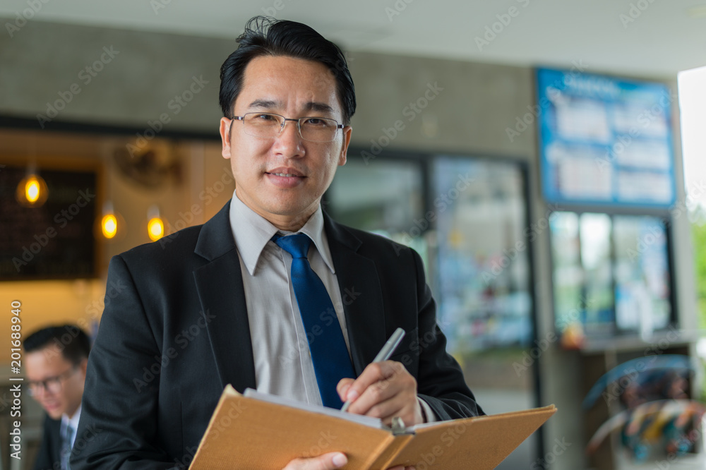 Wall mural Asian businessman holding a book and pen to write a business plan. He standing and looking forward, thinking for business future and financial success.