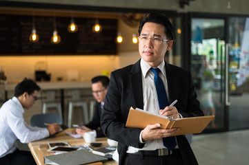 Asian businessman holding a book and pen to write a business plan. He standing and looking forward, thinking for business future and financial success.