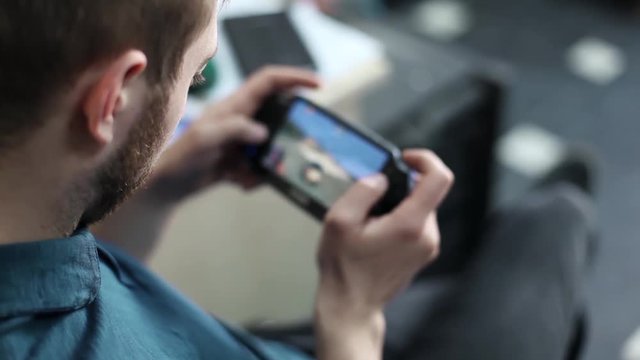 Young Man is playing in a portable game console sitting on a chair at home. Focus is on the man, background blur. Man using console.
