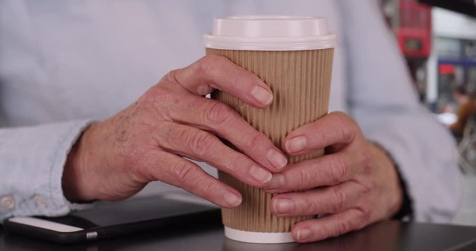 Tight shot of caucasian senior hands holding hot beverage cup outside in city, Close up of elderly person's hands grabbing coffee cup on outdoor cafe table, 4k