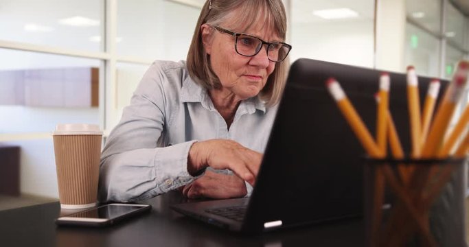 Close view of senior woman with laptop working in office setting indoors, Portrait of elderly woman using computer while sitting inside at the workplace, 4k