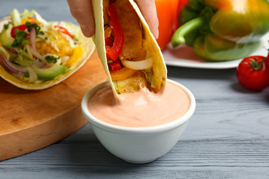 Woman dipping fish taco in sauceboat with tasty creamy tomato sauce on wooden table