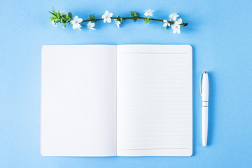 An open blank notebook with a white pen and blossoming branch on a blue background. Spring workplace concept