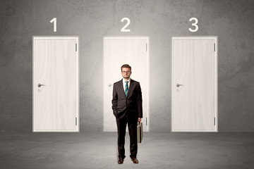 Obraz premium Businessman in grey room with three numbered white door behind his back 