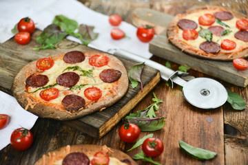 Mini Pizza With Cherry Tomatoes And Salami Sausages On A Wooden Background Natural Rustic, A Pizza Cutter And Ingridienty.