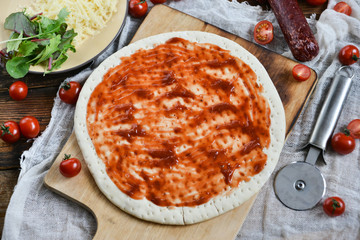 Ingredients for Pizza: basis dough, salami sausage, cherry tomatoes, grated cheese, tomato paste, herbs and arugula. Natural Wooden Background With Linen Napkin and Knife For Pizza