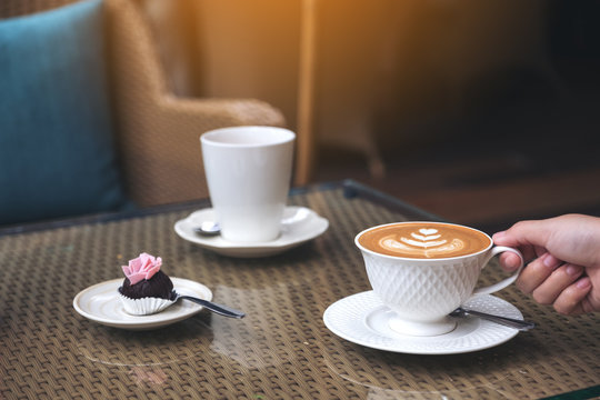 Closeup image of a hand holding a cup of hot latte coffee with latte art and snack on table in cafe