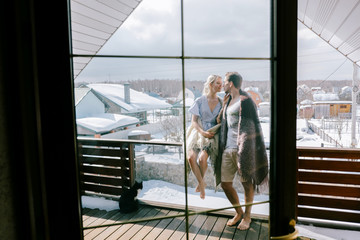 Lovely couple together at home. Pretty woman and attractive man wearing stylish casual clothes and blanket standing on a balcony and hugging