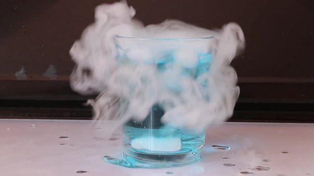 Dry ice in a glass of blue water and engender white smoke on white floor with black background.